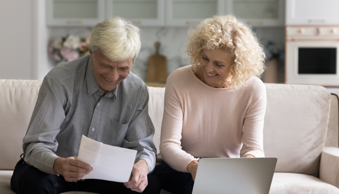 Older couple working on laptop at home smiling while reviewing paperwork.