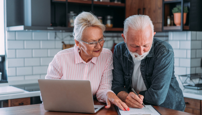 Older couple reading paperwork and using laptop at home.